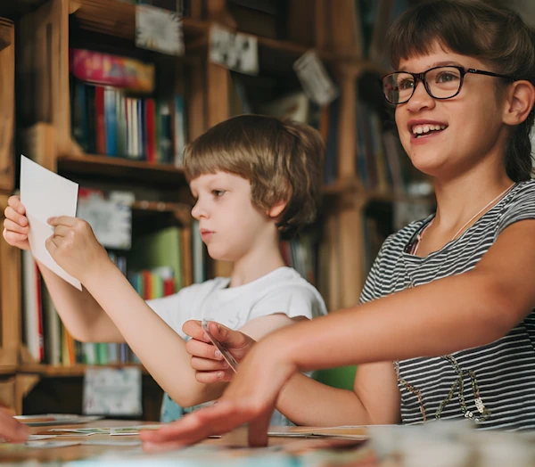 Двое детей на занятиях в чемодане сказок. Девочка на переднем плане улыбается и прижимает что-то рукой к столу.