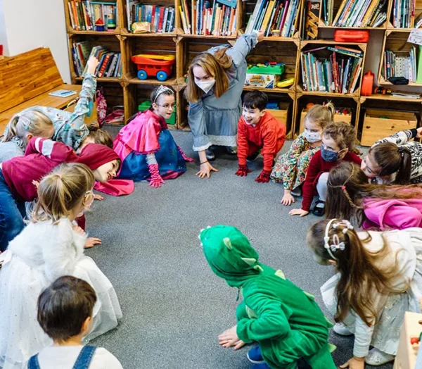 Множество детей и руководитель сидят на полу в игровой комнате. Они расположились в круг упёршись руками об пол.
