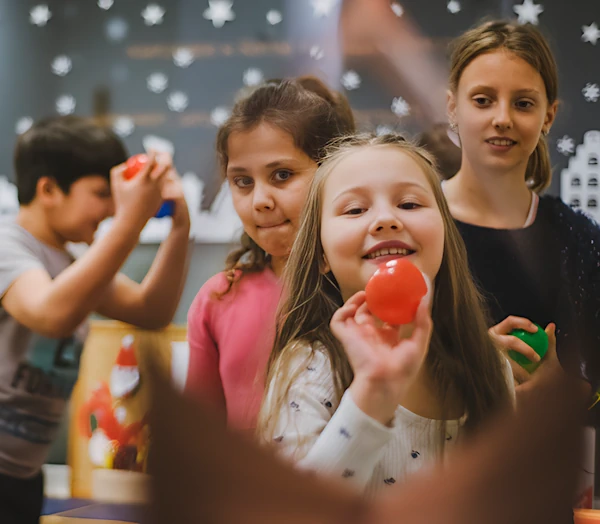 Девочка, держа в руке не большой красный мяч хочет бросить его в сторону камеры. Другие дети наблюдают за ней.