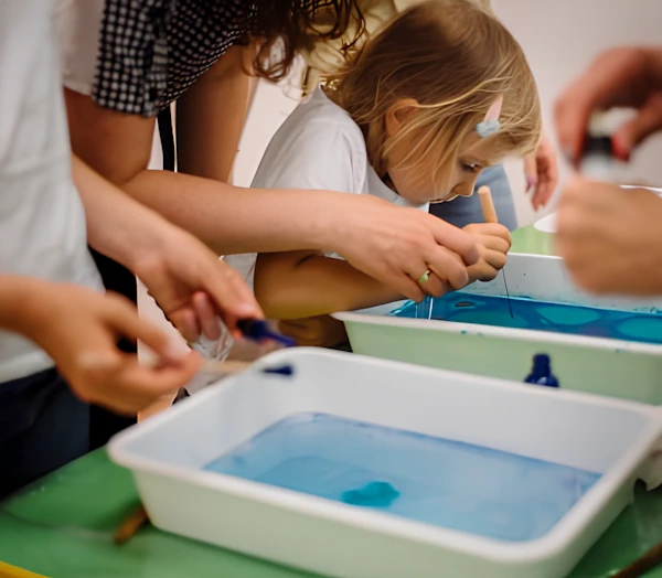 Девочка на занятии смешивает краску в ванночке с водой. Преподаватель, держа за руку, помогает её.