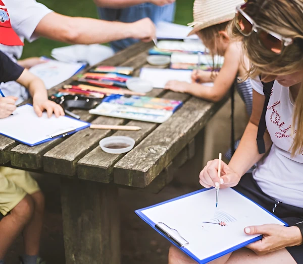 Семья на уличном мастер-классе по рисованию делает эскизы сидя за деревянным столом.