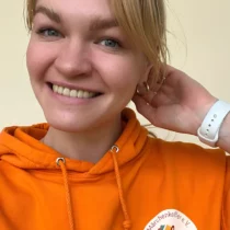 Девушка в оранжевой толстовке с логотипом организации, на руке часы, улыбается.