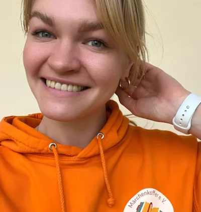 Девушка в оранжевой толстовке с логотипом организации, на руке часы, улыбается.